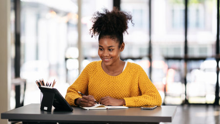 African american student woman in sweater watching e-learning to studying lesson online class on tablet and taking notes in notebook with pencil while learning knowledge and education in university.の写真素材