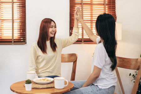 Young woman lesbian couple touching hand to giving high five for celebration after receives good news while spending time to relaxation together in living room at home.の写真素材