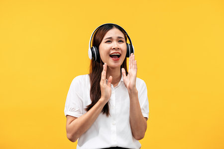 Young asian woman wearing headphone and white short sleeve shirt while listening music and singing the song isolated over yellow background.の写真素材