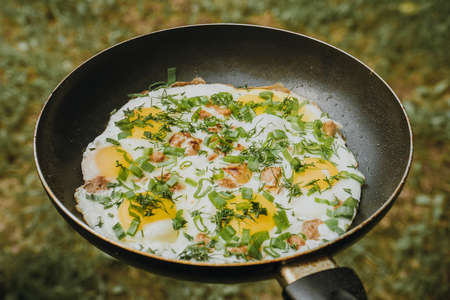 Frying pan of fried eggs, bacon and organic greens, green grass on the background, cooked on the grill on a summer sunny day, summer breakfast, eating in nature - the concept of a healthy lifestyle. High quality photoの写真素材