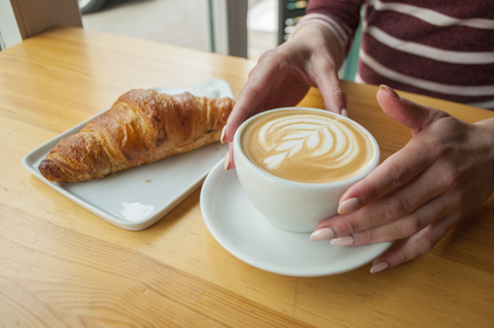 Young lady's hands with beautiful manicure holding a cup of coffee with a croissant on the wooden table.の写真素材