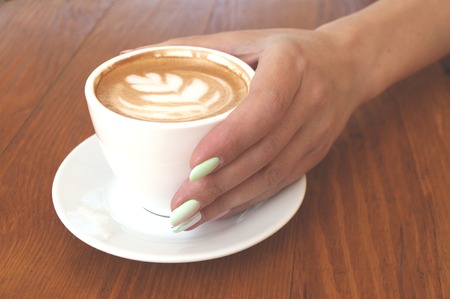Young lady's hand with beautiful manicure holding a cup of cappuccino with latte art on wooden table in a cafe.の写真素材