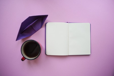 Paper ship, a cup of morning coffee and notebook of violet color on pastel pink background with text space.の写真素材