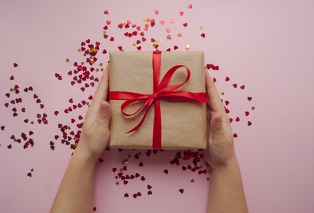 Child is holding gift box wrapped in brown colored craft paper and tied with red bow on pink...