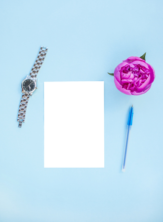 Flat lay of beautiful pink peony flower on blue background with pen, watches and white isolated paper.の写真素材