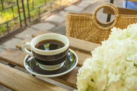 A cup of black coffee on a wooden table and a straw bag with hydragea flowers on the background.