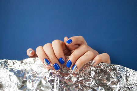 Young woman's hands with beautiful manicure blue on blue background with metallic shiny paper. 2020...