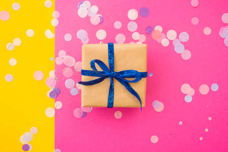 Giftbox tied with blue color ribbon on bright pink, pastel yellow background with colorful confetti...