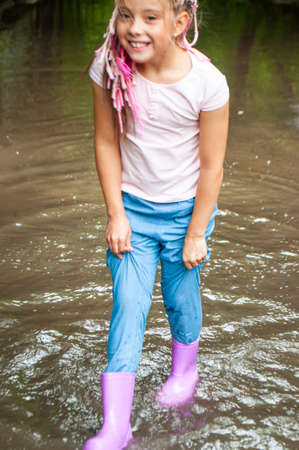 A happy braided girl in rubber boots is playing in muddy puddles.