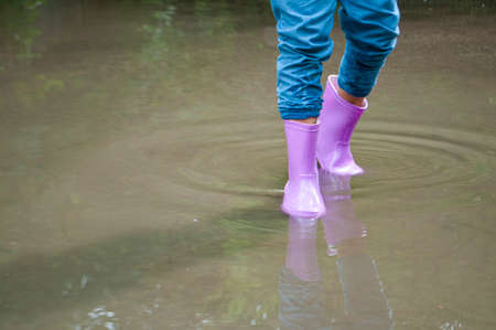 A girl in purple rubber boots is playing in muddy puddles.