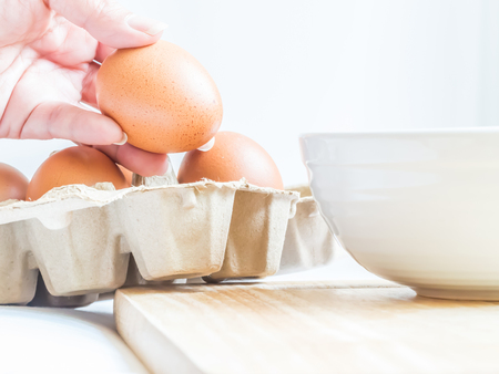 Eggs in paper tray with woman hand, wooden cutting board and white bowl for preparing meal.
