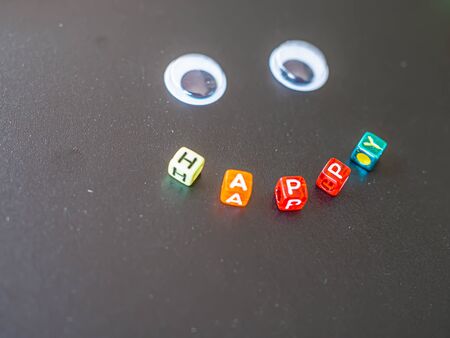 Closeup of multicolored alphabet plastic box of happy word in smile shape on black board with blurry eyes toy. Happiness concepts and ideas.の写真素材