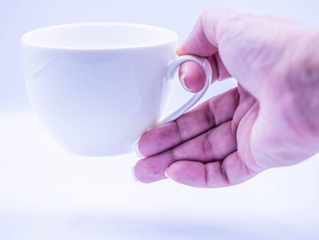 Closeup of woman's hand holding or handle white ceramic coffee or tea cup on gray floor and wall background in the kitchen, living room or cafe for drinking.の写真素材