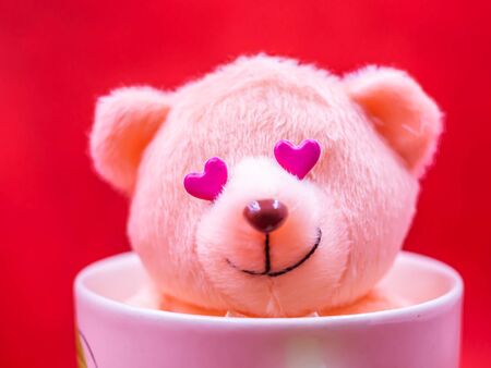 Closeup of pink heart shape sticker,  smiling teddy bear doll in white coffee mug on vivid, vibrant red background for happiness, cheerful, love, valentine, positive thinking concepts and ideas.の写真素材