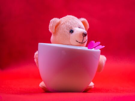 Closeup of smiling teddy bear doll with pink heart shape sticker and white coffee cup on vivid, vibrant red background for happiness, cheerful, love, valentine, enjoyment concepts and ideas.の写真素材