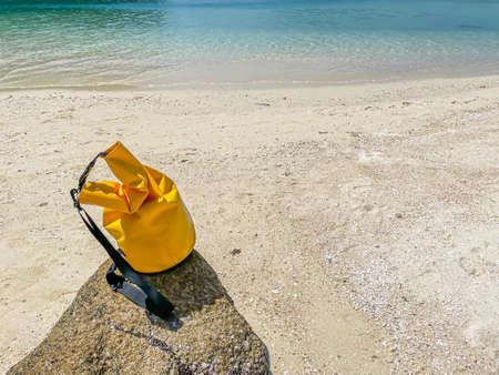 Closeup of vivid yellow waterproof bag on the rock or stone with blurry beach, and sea background. Summer or travel concept and idea.の写真素材