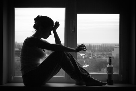 Sad young woman sitting on the window, drinks wine. Alcoholism problem. Black and white photography.の写真素材