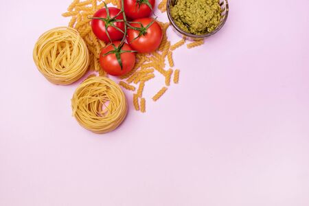 Variety of Types and Shapes of Dry Pasta Tagliatelle Fusilli Raw Tomatoes Bowl of Pesto Top View Pink Background Copy Space Italian Foodの写真素材