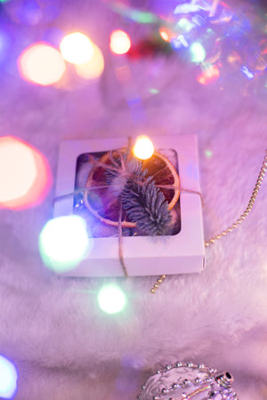 A small Christmas gift box decorated with dried orange and fir branch, laying on fur cloth, led neon lights and tree behindの写真素材