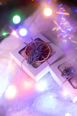 A small Christmas gift box decorated with dried orange and fir branch, laying on fur cloth, led neon lights and tree behindの写真素材
