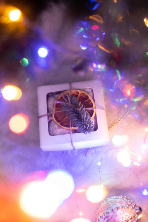 A small Christmas gift box decorated with dried orange and fir branch, laying on fur cloth, led neon lights and tree behindの写真素材