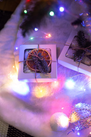A small Christmas gift box decorated with dried orange and fir branch, laying on fur cloth, led neon lights and tree behindの写真素材