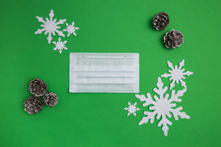 Christmas themed flat lay shot with medical mask on green background with snowflakes and fir cones aroundの写真素材