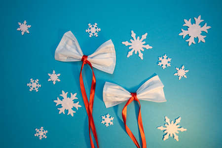Two bows made of medical masks and red ribbons on blue backdrop with white snowflakesの写真素材