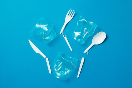 White broken single-use plastic knives, spoons, forks and plastic drink straws on a blue background....