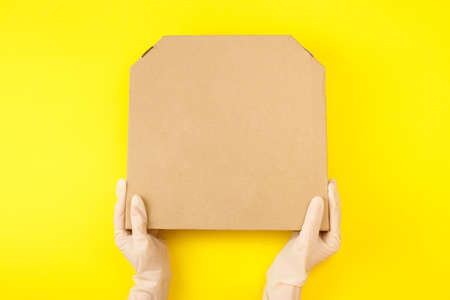 Hands in protective gloves hold a pizza box on a yellow background. Food delivery and virus protection conceptの写真素材
