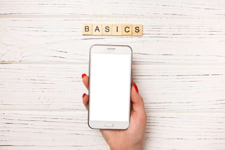 Basic income written on wood blocks with woman hand and smartphone on white wooden background. Back to basics fundamental principles conceptの写真素材