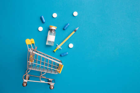 Flat lay supermarket trolley, vaccine and syringe on a blue background. Vaccination of people, prevention of coronavirus epidation. Concept of buying a vaccineの写真素材