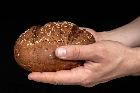 Man's hands hold tasty fresh loaf of dark bread with sesame seeds on black background.