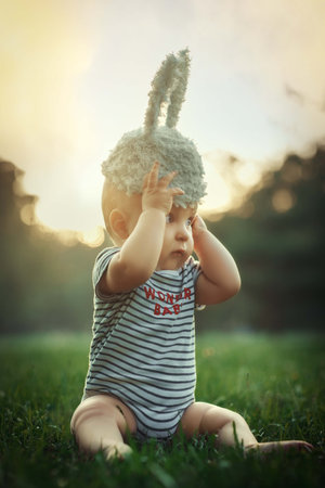 Cute baby boy in bunny ears sitting on green grass at sunsetの写真素材
