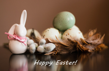 Small white rabbit figurine with easter eggs, willow branch and feathers on pastel background with text Happy Easterの写真素材