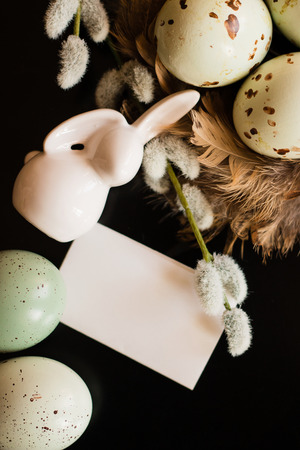 Small white rabbit figurine with easter eggs, willow branch and feathers' nest on black background. Holiday background.View from above.の写真素材