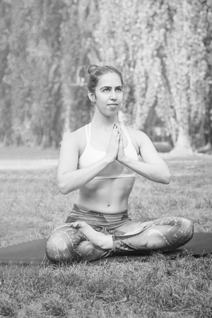 âYoung woman practicing yoga in nature. Sitting on grass and meditating, holding hands together. Healthy and sport concept.  Copy space for text. Black and white photo.の写真素材