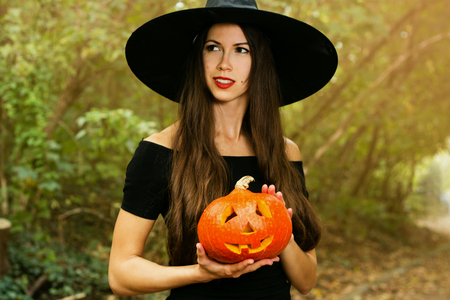 Halloween holiday background. Halloween Witch with a halloween pumpkin jack o lantern decor with funny face in a dark forest. Beautiful young woman in witches hat and costume holding pumpkin.の写真素材