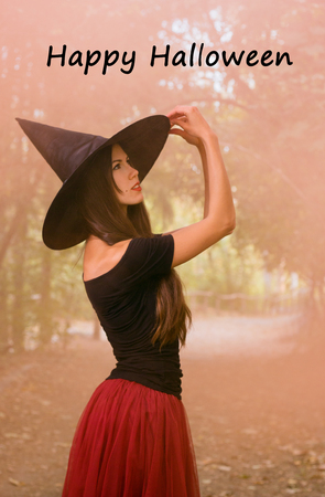 Halloween holiday background. Halloween Witch in a dark forest. Beautiful young woman in witches hat and costume on orange smoke background with text happy halloween.の写真素材