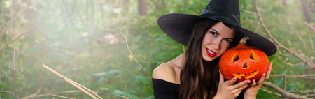 Halloween holiday background. Halloween Witch with a halloween pumpkin jack o lantern decor with funny face in a dark forest with toned effect. Beautiful young woman in witches hat and costume holding pumpkin.の写真素材