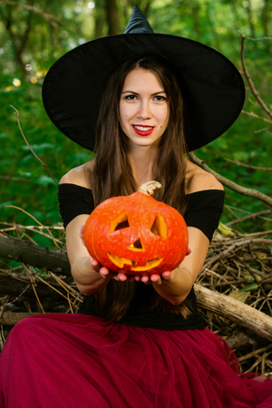 Halloween holiday background. Halloween Witch with a halloween pumpkin jack o lantern decor with funny face in a dark forest. Beautiful young woman in witches hat and costume holding pumpkin.の写真素材