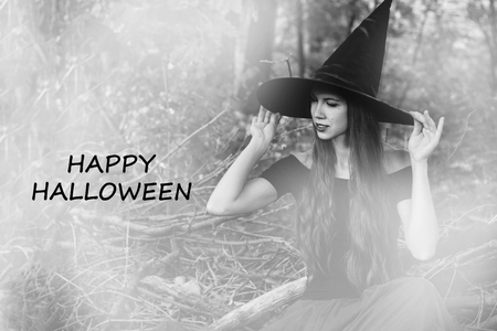 Halloween holiday background. Halloween Witch in a dark forest with toned effect. Beautiful young woman in witches hat and costume with text happy halloween. Black and white photo.の写真素材