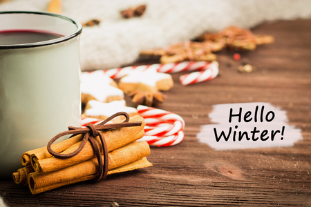 Winter theme. Hot steaming cup of glint wine with spices, cinnamon, anise, cookies in a shape of star, red candies, pepper and gray scarf on wooden background with text Hello Winter. View from above.の写真素材