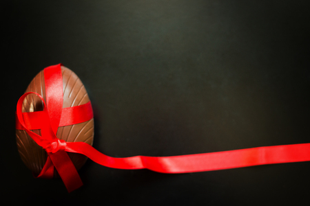 Big chocolate Easter egg decorated with red ribbon on black background. Holiday Easter background with copy space for text. Top view.の写真素材