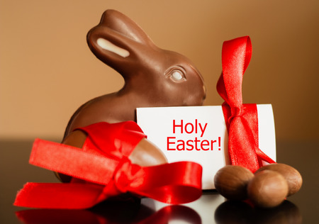 Big and small chocolate Easter eggs, traditional Easter chocolate Bunny decorated with red ribbon on pastel background. Holiday Easter background with text Holy Easter.の写真素材