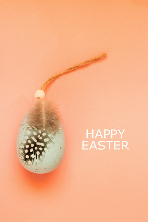 Blue pastel big Easter egg decorated with ribbon and bird feather on coral background. Holiday Easter background with text Happy Easter. Top view.の写真素材