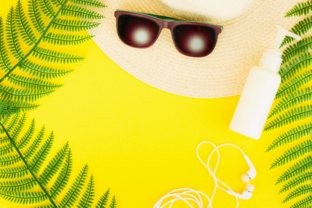 Summer, travel, vacation concept flat lay. Beach accessories: sunscreen lotion, summer hat, sunglasses, fern and white headphones on yellow background. Top view. Space for text.の写真素材