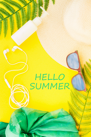 Summer, travel, vacation concept flat lay. Beach accessories: sunscreen lotion, summer hat, sunglasses, shawl, fern, and white headphones on yellow background. Top view with text hello Summer.の写真素材