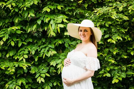 Pregnancy, love, people and expectation concept - happy pregnant woman posing over green natural background in white dress and hat, holding belly with both hands.の写真素材
