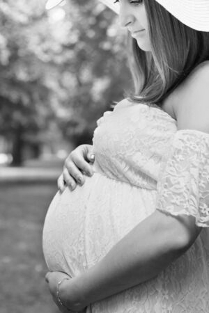 Pregnancy, love, people and expectation concept - happy pregnant woman posing over green natural background in white dress, holding belly with both hands. Black and white photo.の写真素材